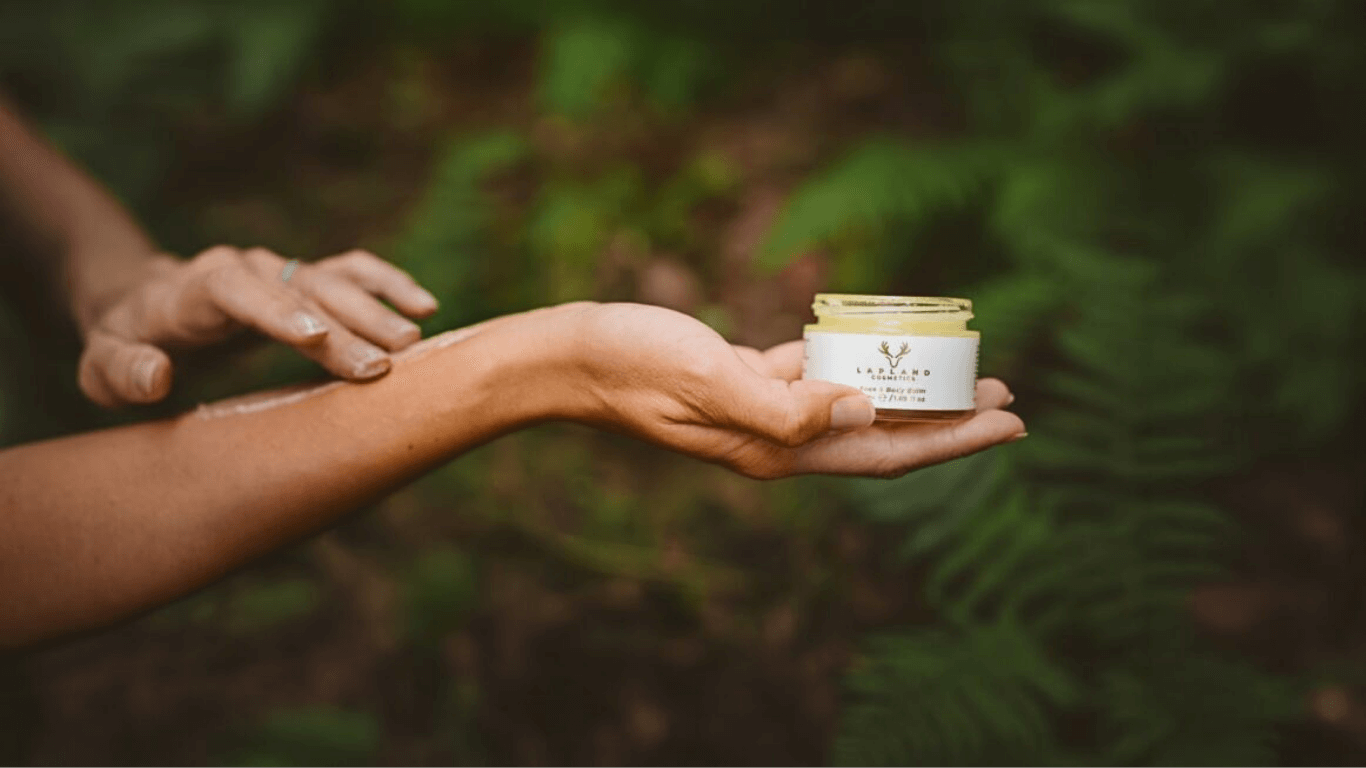 Ženské ruce s tělovým balzámem, jedna na druhou nanáší balzám, na pozadí lesa.